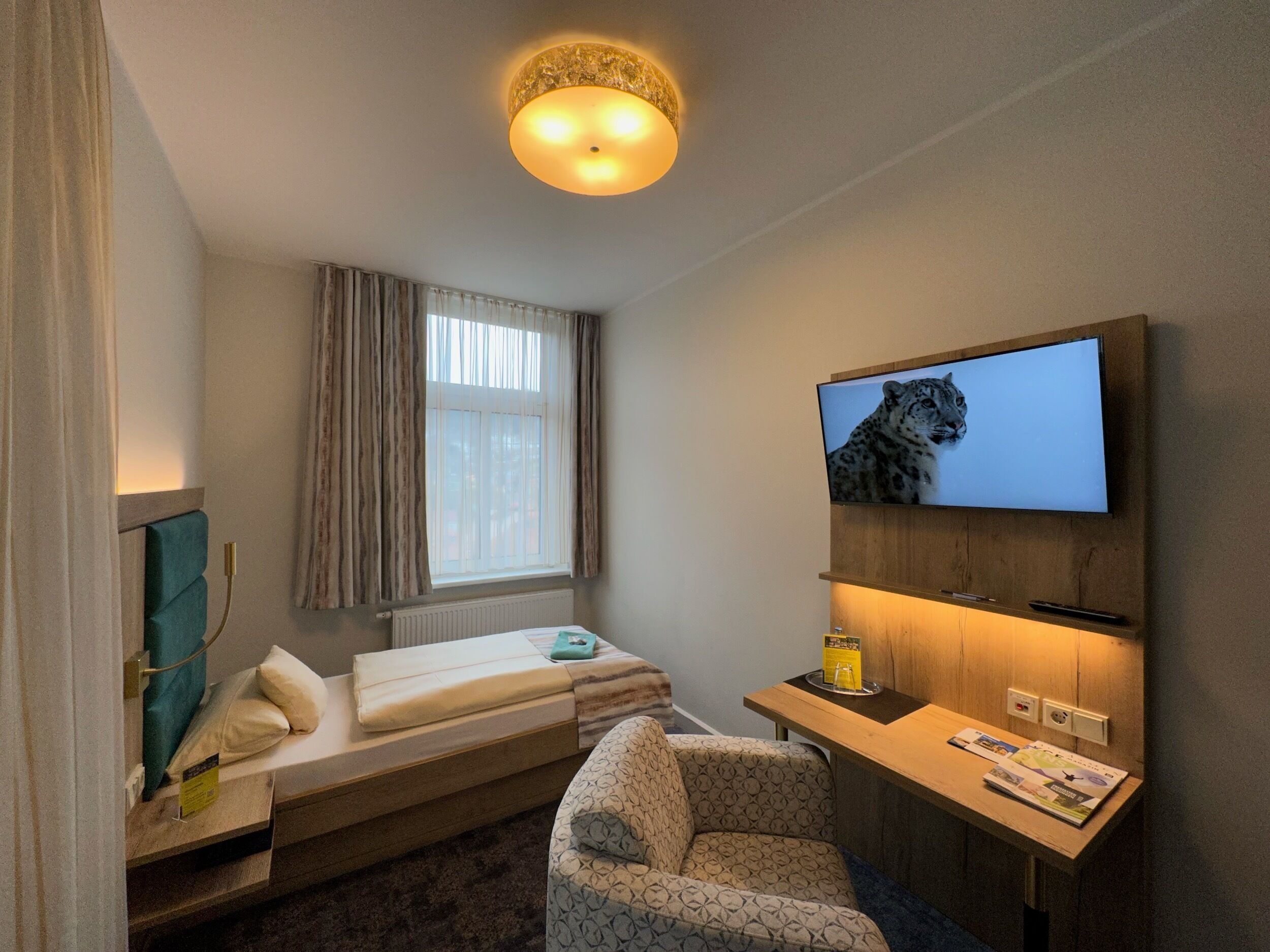 A compact hotel room with a bed, a desk, a chair, a television, and a window with curtains.