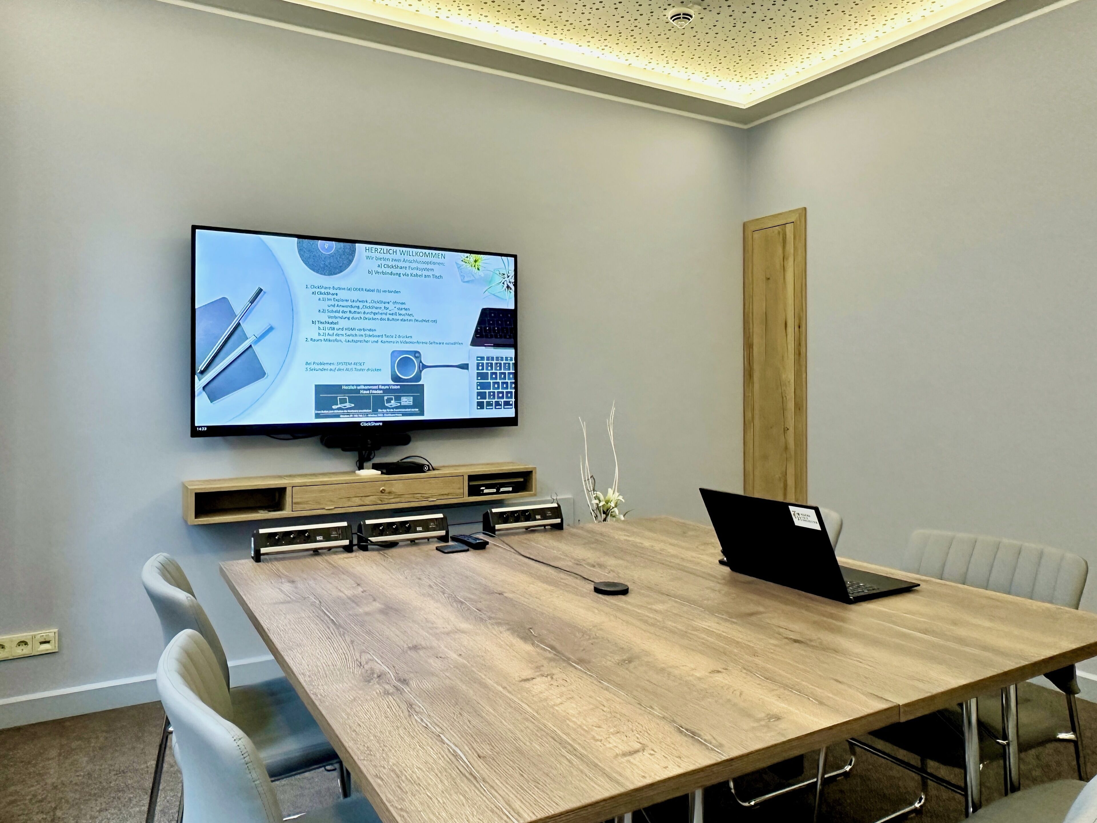 A conference room with a large flat-screen TV mounted on the wall, a wooden table, and several chairs.