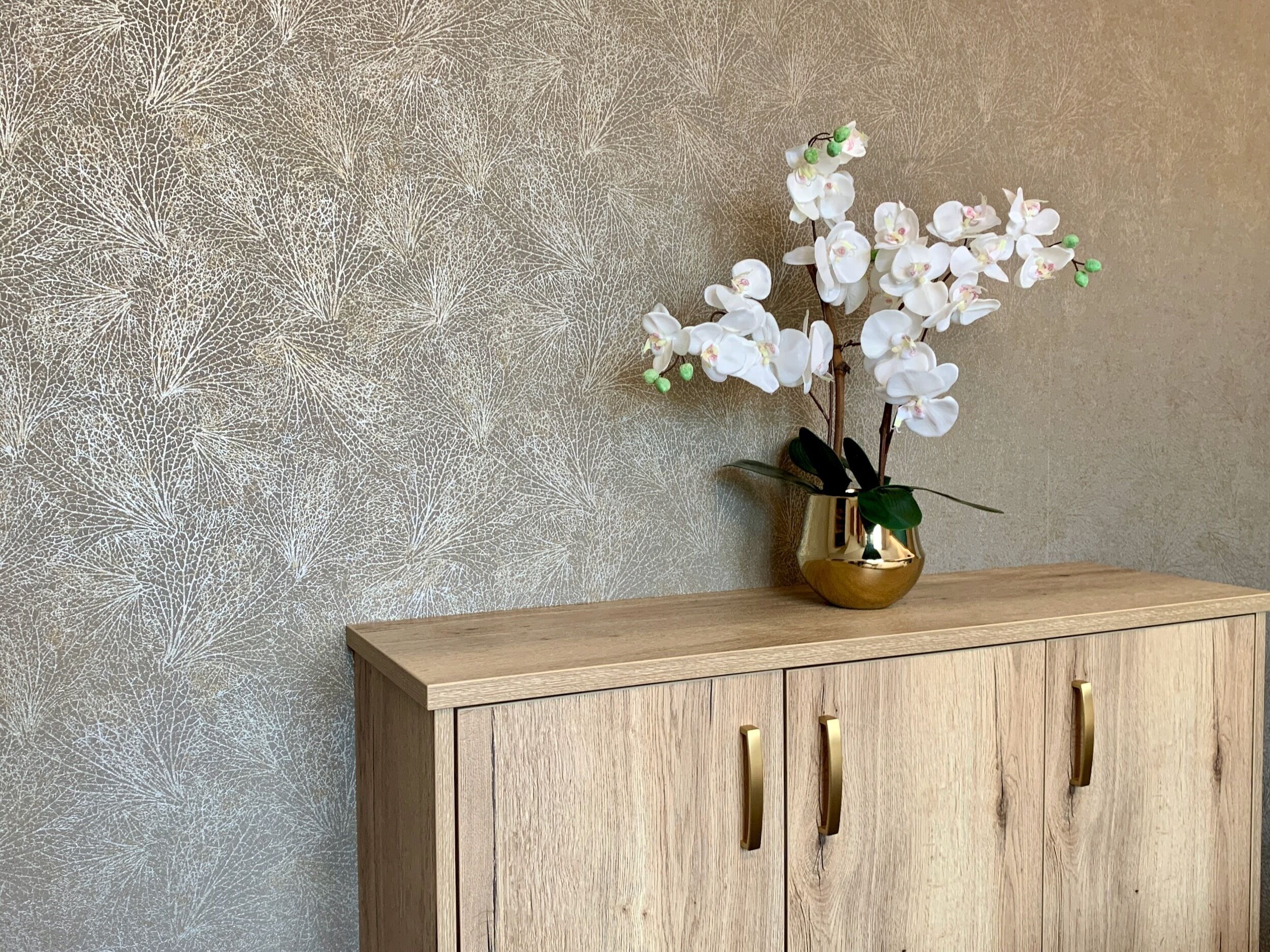 Et træskab med messinggreb og en guldfarvet vase med hvide blomster.