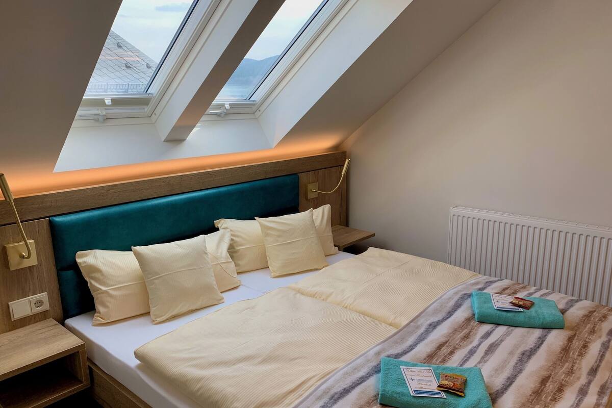 Un dormitorio con tragaluces, una cama con ropa de cama blanca, cabecera color aguamarina y una mesita de noche con lámpara.