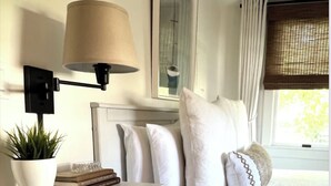 A neatly arranged bedroom with a white bed, a bedside table with a lamp, a framed picture above the bed, and a window with curtains. - Prime Location, Top Floor 1-Bedroom, 1-Bath Condo (Provincetown)