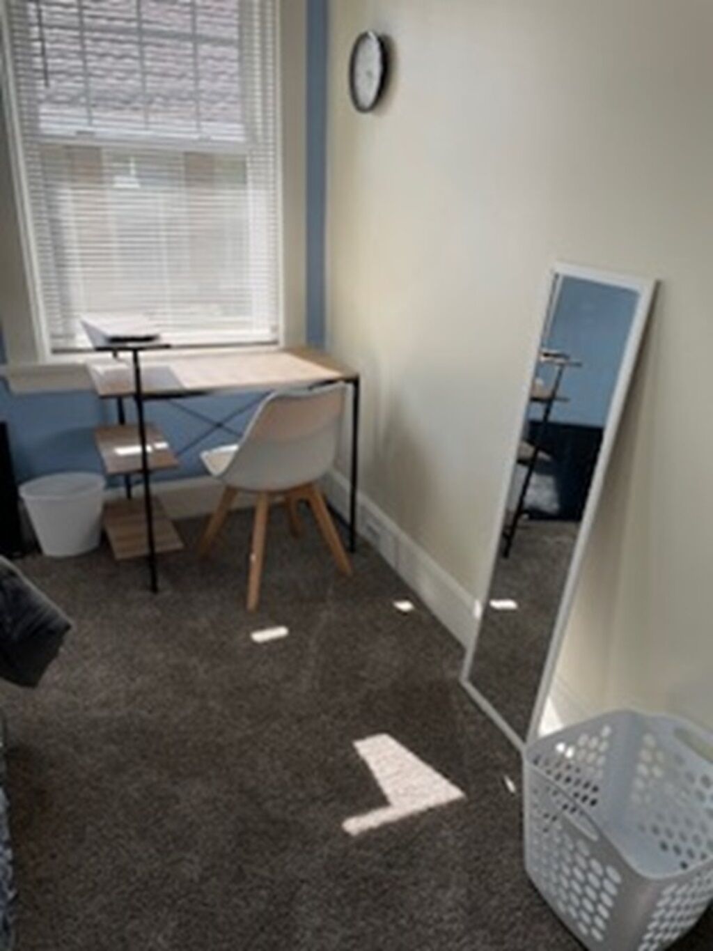 A room with a desk, chair, window with blinds, a clock, and a mirror on the wall.