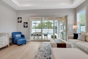 Una sala moderna con un sillón azul, un sofá beige, una mesa de centro y vistas a un área exterior con piscina a través de puertas de vidrio.