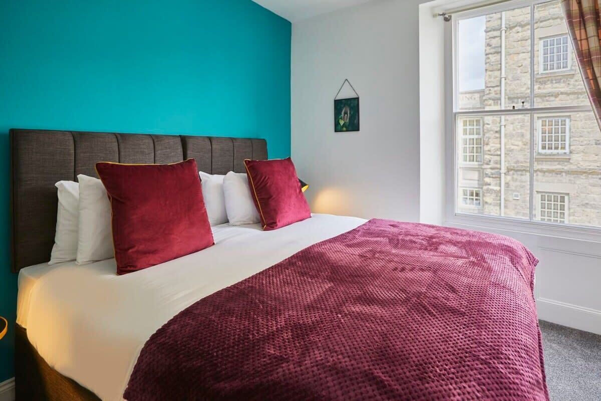 Una cama con cabecera oscura, almohadas blancas y rojas, y una manta rosa en una habitación con paredes color turquesa y una ventana con vistas a un edificio de piedra.