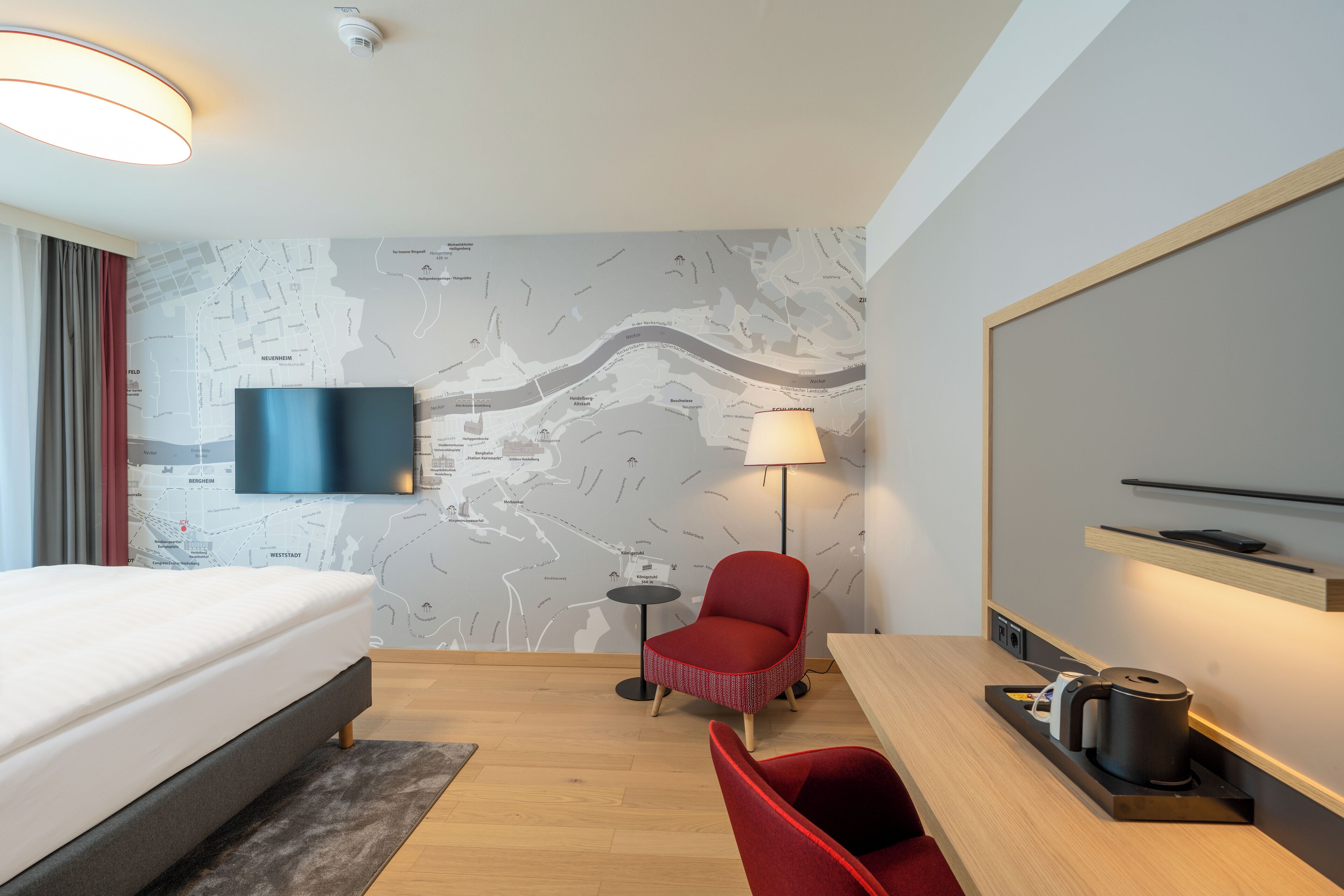 A hotel room with a bed, a desk with a kettle, a red chair, a map on the wall, and a TV.