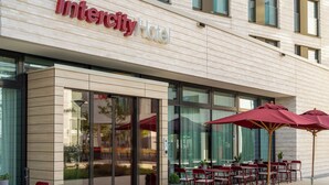 Esterno di un Intercity Hotel con facciata moderna, ampie vetrate e area salotto all'aperto con ombrelloni rossi.