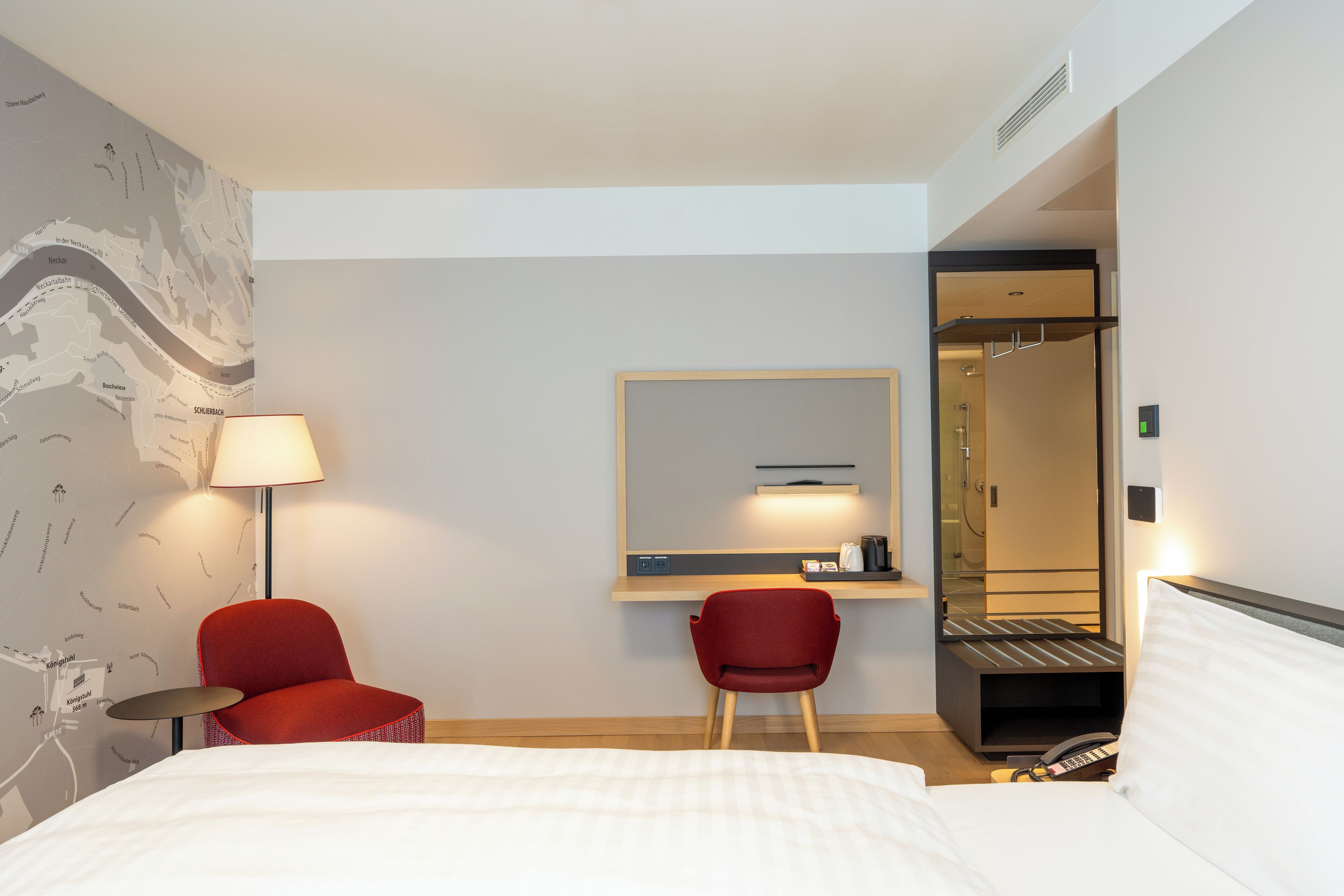 Une chambre d’hôtel avec un lit, deux chaises rouges, un bureau avec une lampe, un miroir et un espace douche.
