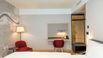 Une chambre d’hôtel avec un lit, deux chaises rouges, un bureau avec une lampe, un miroir et un espace douche.