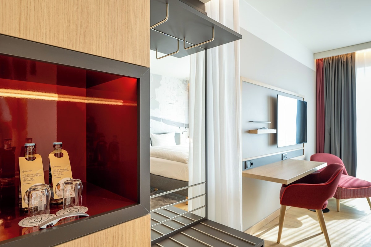A modern hotel room with a wooden desk, red chairs, a TV, and a minibar with bottles and glasses.