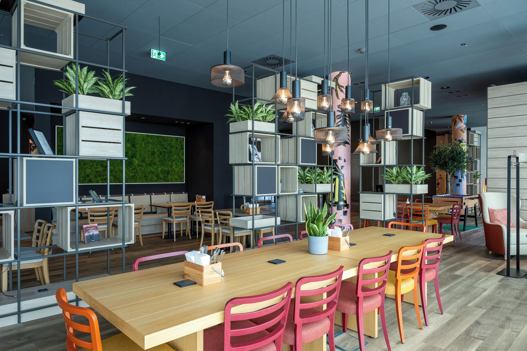 Un comedor moderno con una larga mesa de madera, sillas rojas y lámparas colgantes. Hay plantas en macetas y estanterías con libros y objetos decorativos.