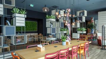 Un espace de salle à manger moderne avec une longue table en bois, des chaises rouges et des lampes suspendues. On y trouve des plantes en pot et des étagères remplies de livres et d’objets décoratifs.