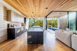 A modern kitchen with a central island, a dining area with a long table, and a pool area visible through large windows.
