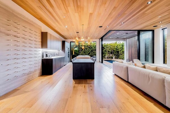 A modern interior with a wooden ceiling, a central dining area, and a seating area with a view of the outdoors.