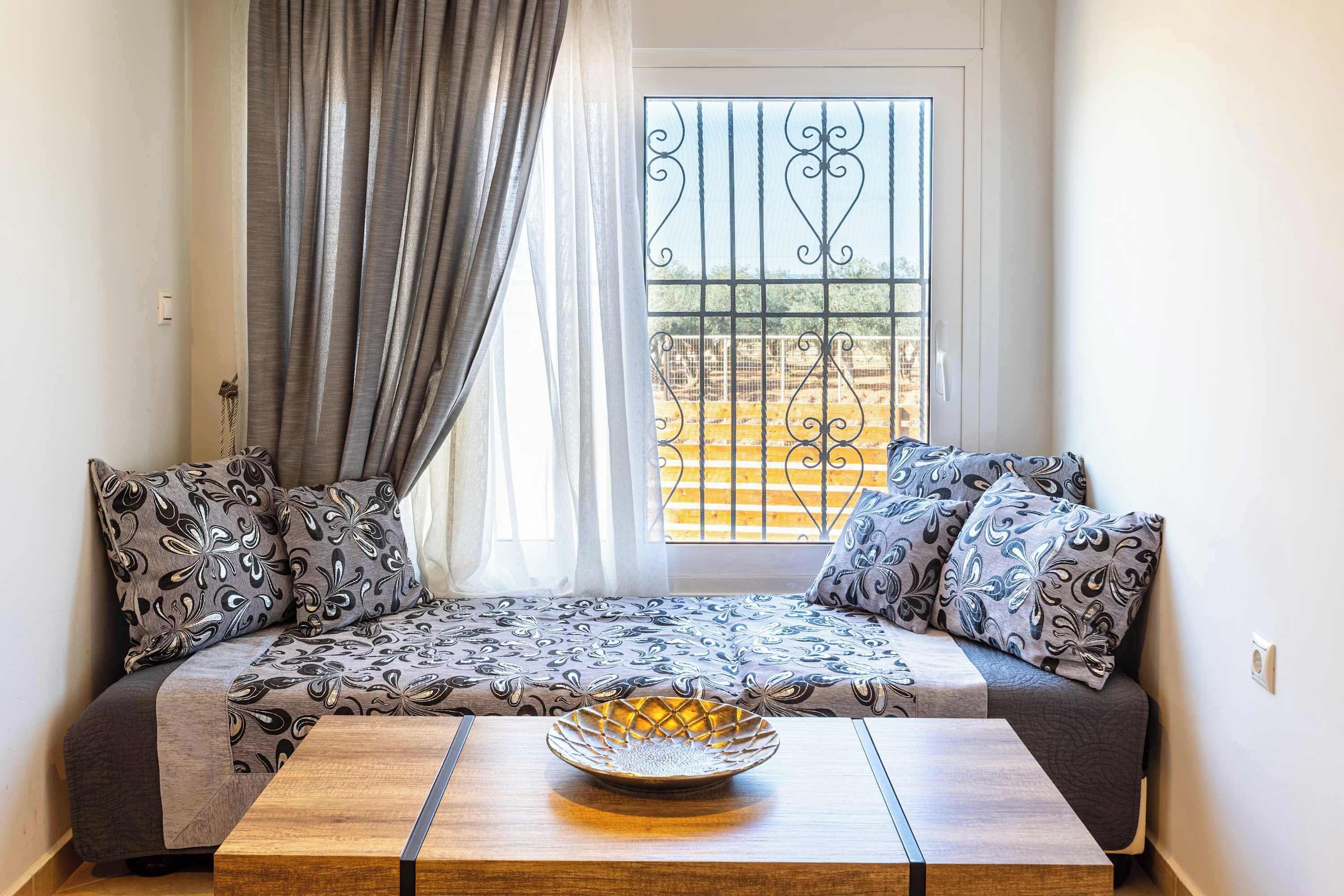 A cozy seating area with patterned cushions, a wooden table with a decorative bowl, and a view through a window with metal bars.