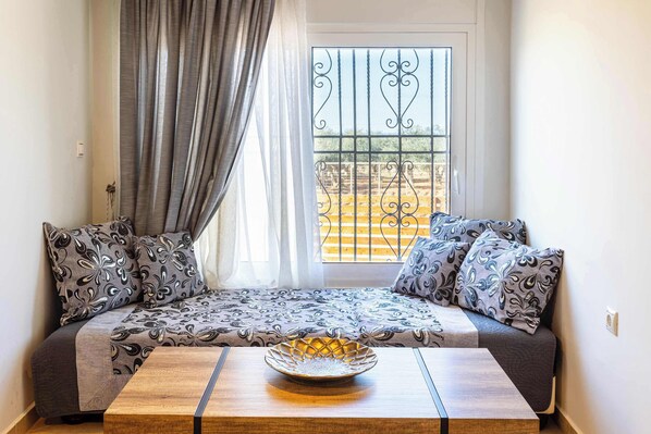 A cozy seating area with patterned cushions, a wooden table with a decorative bowl, and a view through a window with metal bars.