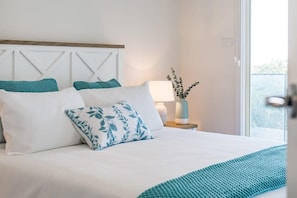 A neatly made bed with white and teal bedding, a bedside lamp, and a view of the outdoors.