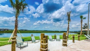 Un área de descanso junto a la playa con sillas y vistas a un lago, rodeada de palmeras y un cielo despejado.