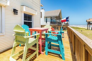 A wooden deck with colorful Adirondack chairs and a small table, overlooking a beach and a house with a balcony. - 2nd Row Beachside w/ Gulf Views, Updated Interior, 2 Decks & Easy Beach Access (Galveston)