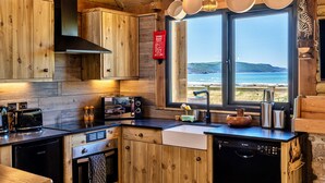 A wooden cabin kitchen with a view of the sea, a fire extinguisher, and modern appliances. - Driftwood (Widemouth Bay)