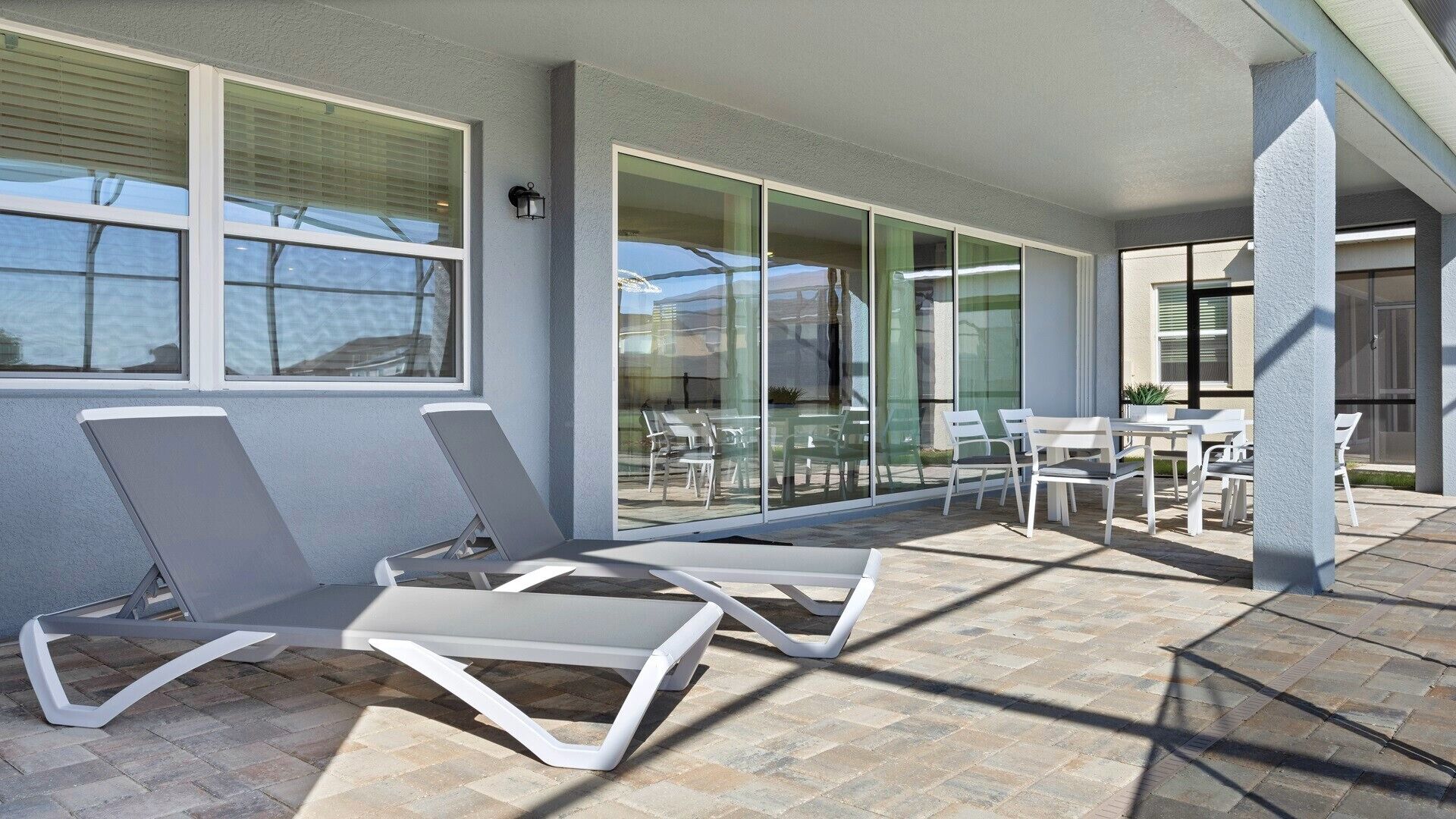 En moderne udendørs terrasse med liggestole, en glasdør der fører til et overdækket område, og en gulvbelagt med natursten.