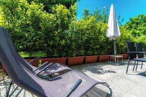 A patio area with lounge chairs and a white umbrella, surrounded by lush green bushes and potted plants. - Casa Mummi (Losone)