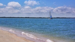 A beach with clear blue water, a sailboat, and a distant shoreline with buildings.