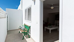Un mur extérieur blanc, un banc en bois, un ventilateur de plafond et un coin repas visible à travers une porte ouverte.