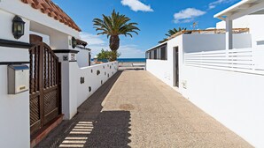 Un étroit sentier mène à un bâtiment blanc surmonté d’un toit en tuiles, avec un palmier et un ciel d’un bleu limpide.