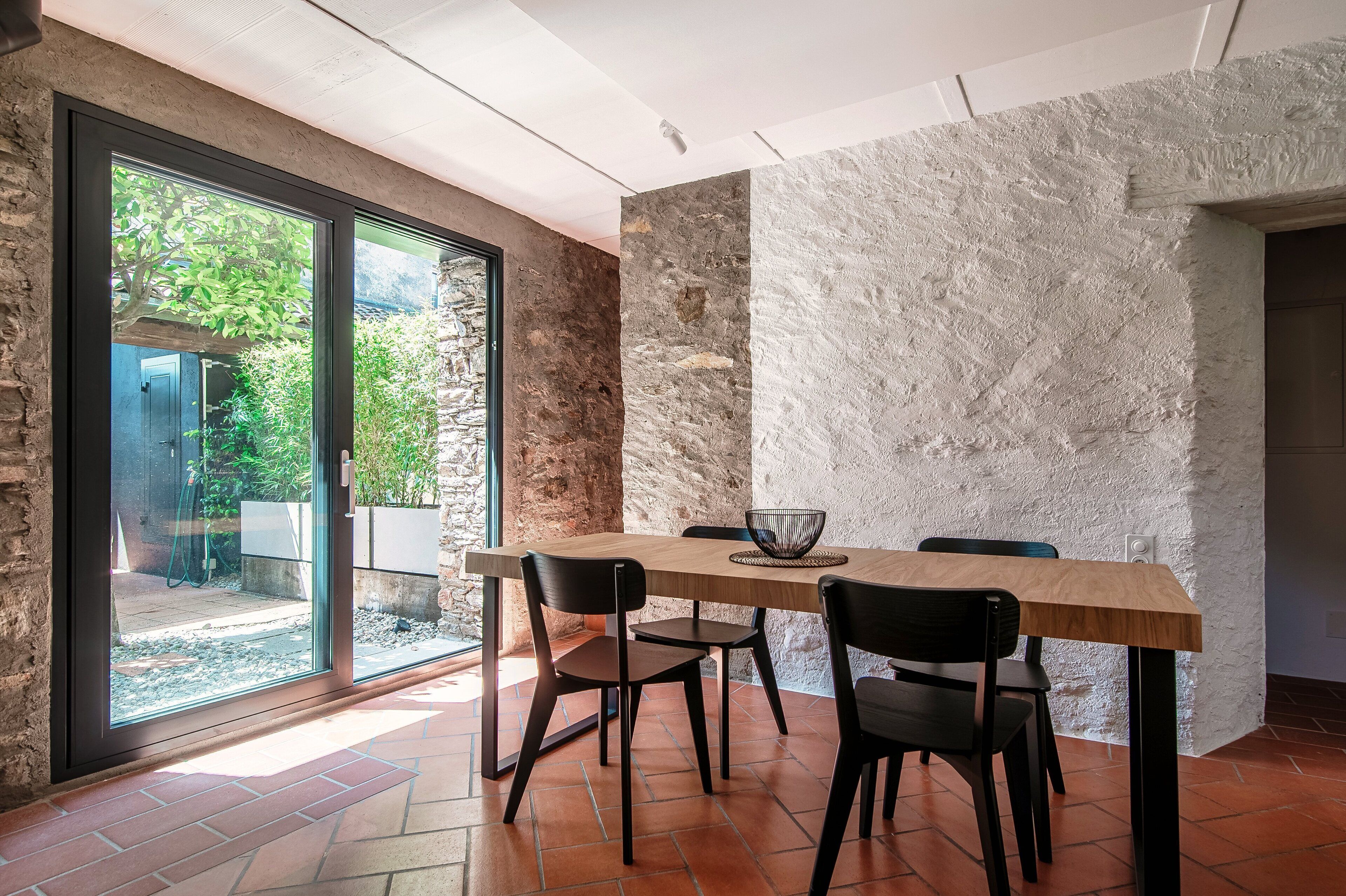 Un área de comedor con una mesa grande y cuatro sillas, una pared de piedra y vistas a un jardín exterior a través de puertas de vidrio.