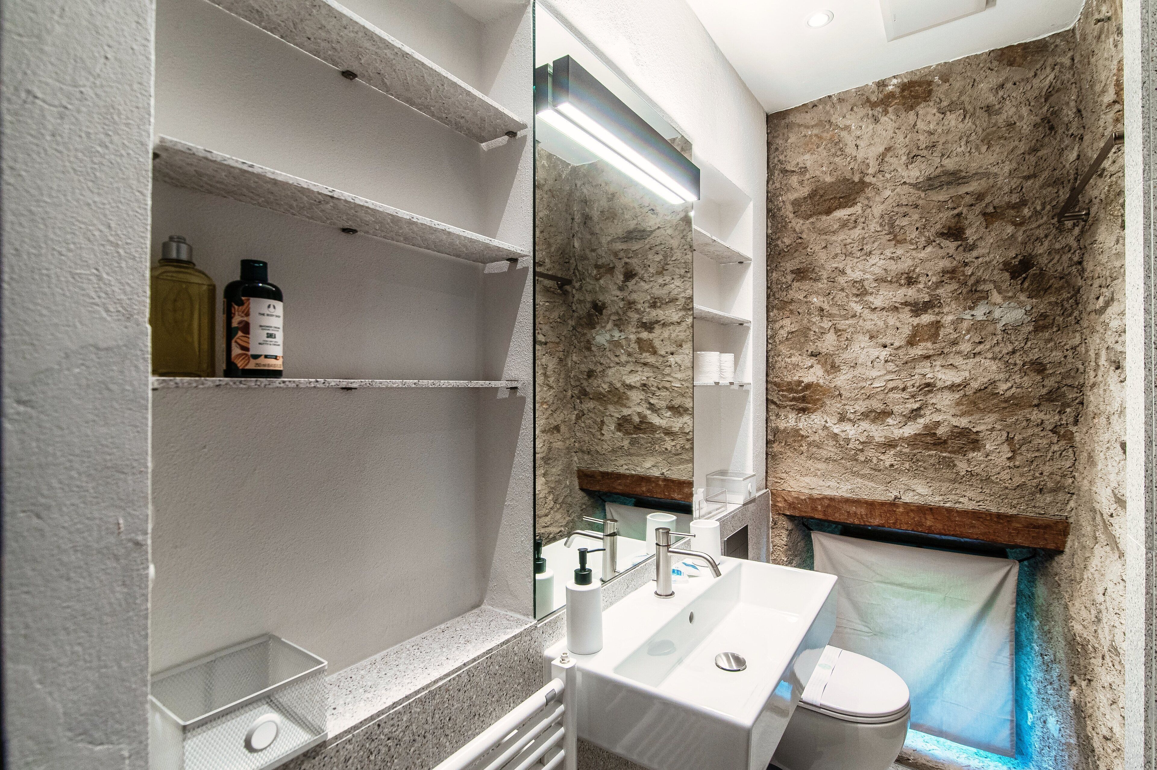 A bathroom with a stone wall, a white sink, a mirror, and shelves with toiletries.