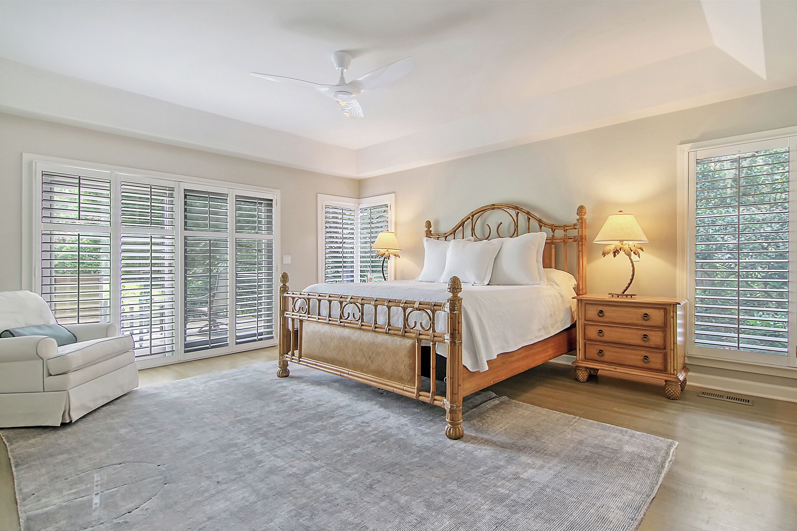 A bedroom with a large bed, two bedside tables with lamps, a ceiling fan, and large windows with blinds.