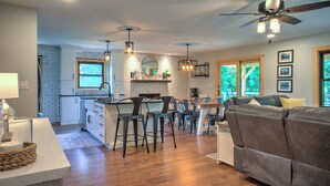 A modern kitchen with white brick walls, wooden flooring, and a central island with bar stools. There is a dining area with a gray sofa and a ceiling fan.