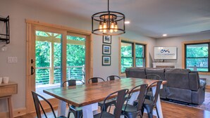 A spacious living area with a wooden dining table, comfortable seating, and large windows offering a view of greenery.