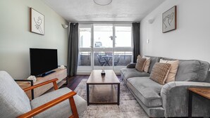 Una sala de estar con un televisor de pantalla plana, un sofá gris, una mesa de centro de madera y un ventanal con cortinas.