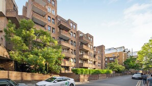 Un edificio residencial de varios pisos con balcones, una calle al frente y coches estacionados a lo largo de un lado.