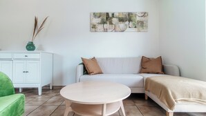 A living room with a white sofa, two brown pillows, a round wooden coffee table, a green armchair, and a white cabinet with a vase of dried plants. - Il Nido di Maria (Losone)