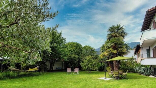 A well-maintained garden with a hammock, two chairs, and a table under a green umbrella, surrounded by lush greenery and a house with a balcony. - Il Nido di Maria (Losone)