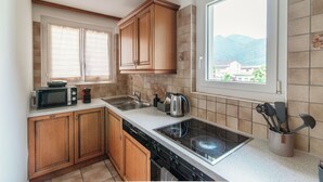 A compact kitchen with wooden cabinets, a microwave, a kettle, and a window with a view of mountains. - Il Nido di Maria (Losone)