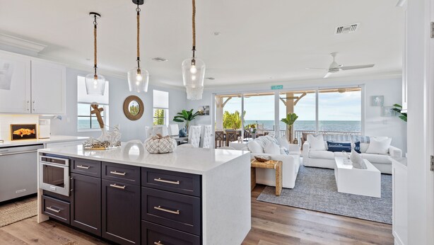 A modern kitchen with dark cabinets and a large island, a dining area with a view of the ocean, and a living room with a white sofa and a coffee table.