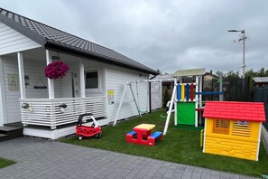 A residential property with a white exterior, a porch with a hanging flower, a small red toy car, a children's play structure, and a yellow playhouse.