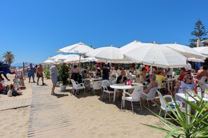 Ett utomhuskafé med vita parasoller och plaststolar, beläget på en sandstrand.