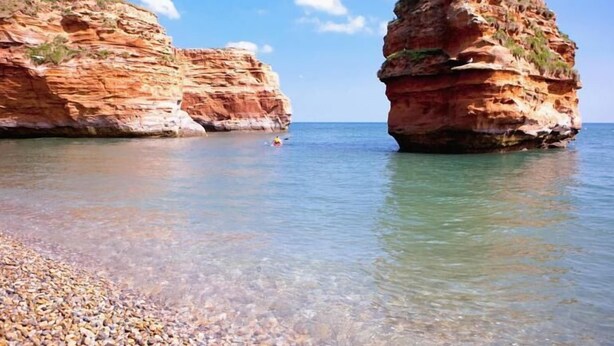 Paesaggio costiero con acque limpide, scogliere rocciose e una spiaggia di ciottoli.