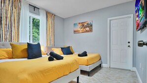 A hotel room with two beds, each with yellow bedding and dark pillows, a white door, a painting of a sunset, and a window with curtains. - Unique Escape GameRoom! Bar, BBQ, Spacious, and Near Beach (Miami)