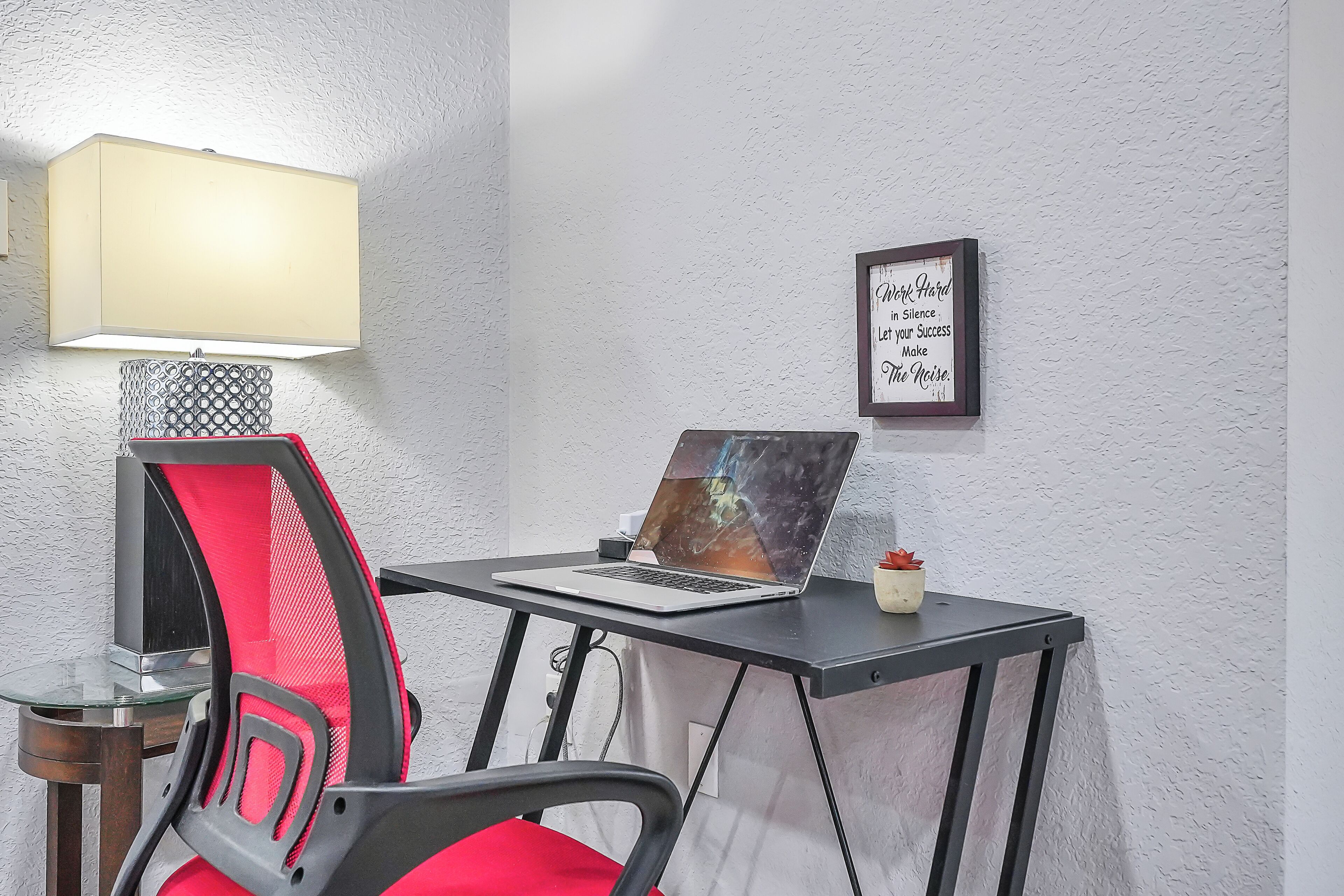 Ein kleiner Arbeitsplatz mit einem Laptop, einer Lampe und einem gerahmten Zitat an der Wand.