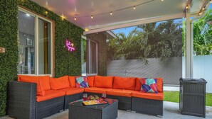 An outdoor seating area with a wicker sofa and a glass coffee table, surrounded by greenery and a clear view of the outdoors. - Unique Escape GameRoom! Bar, BBQ, Spacious, and Near Beach (Miami)