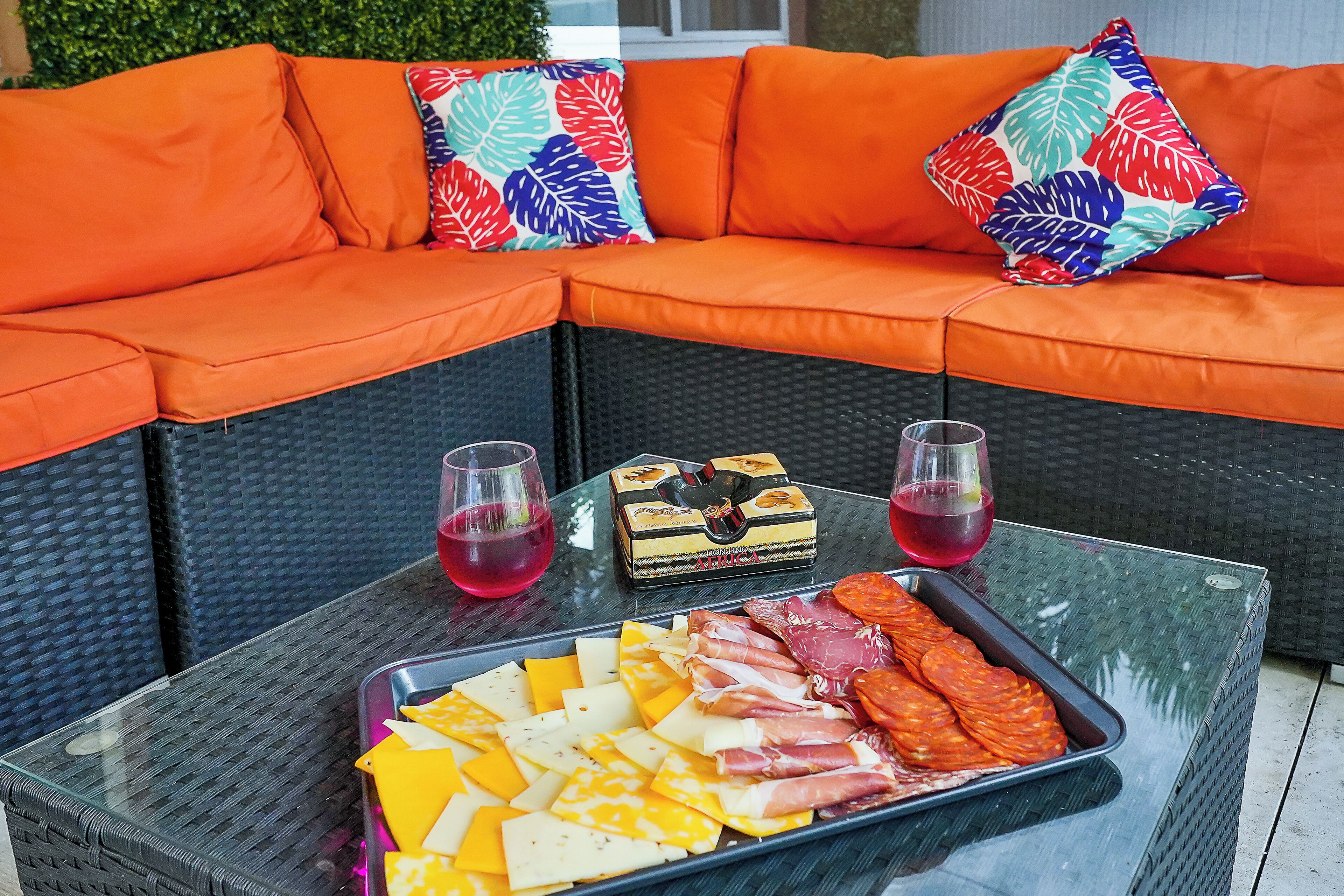 A glass-top table with a charcuterie board, two glasses of red drink, and a wicker sofa with orange cushions and colorful pillows.