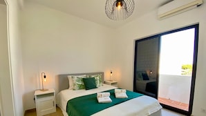 Quarto com cama, mesas de cabeceira, colcha verde e vista para o exterior por uma porta deslizante.