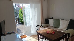 A room with a wooden table, a sofa with cushions, a TV, and a sliding glass door leading to an outdoor area with greenery.