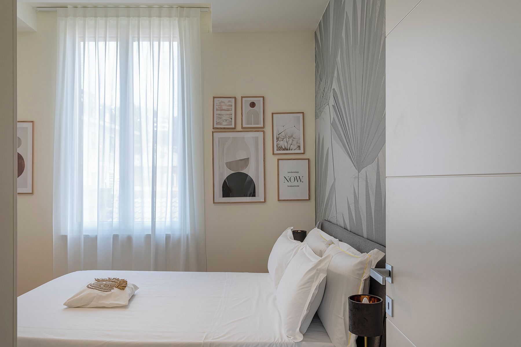 A hotel room with a bed, a bedside table with a lamp, and a window with sheer curtains.