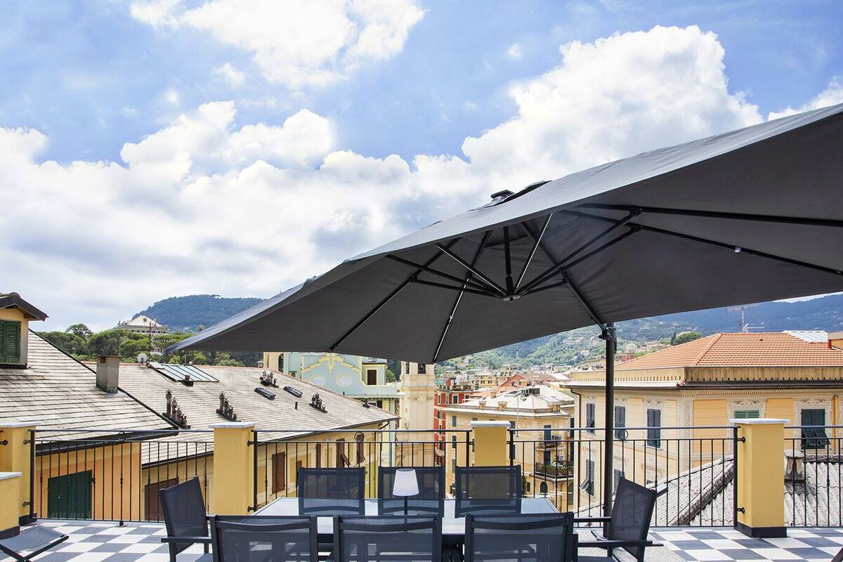 Une terrasse extérieure dotée d’un grand parasol, de tables et de chaises, offrant une vue sur un paysage urbain avec des bâtiments et des montagnes.